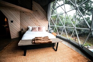 Une chambre à coucher avec un lit, des oreillers, une couverture, un banc en bois et une grande fenêtre donnant sur des arbres.