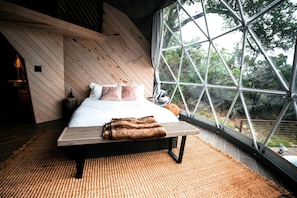 A bedroom with a bed, pillows, a blanket, a wooden bench, and a large window with a view of trees.