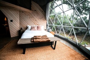 Een slaapkamer met een bed, kussens, een deken, een houten bankje en een groot raam met uitzicht op bomen.