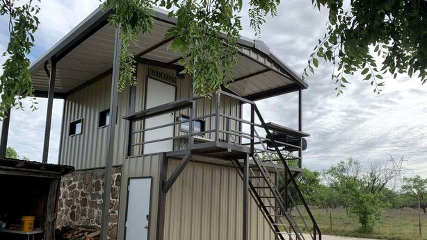 A two-story metal building with a balcony, a stone wall, and a staircase leading to the upper level. - Wilderness Cabin Rental Perfect for Glamping in Texas (Graford)