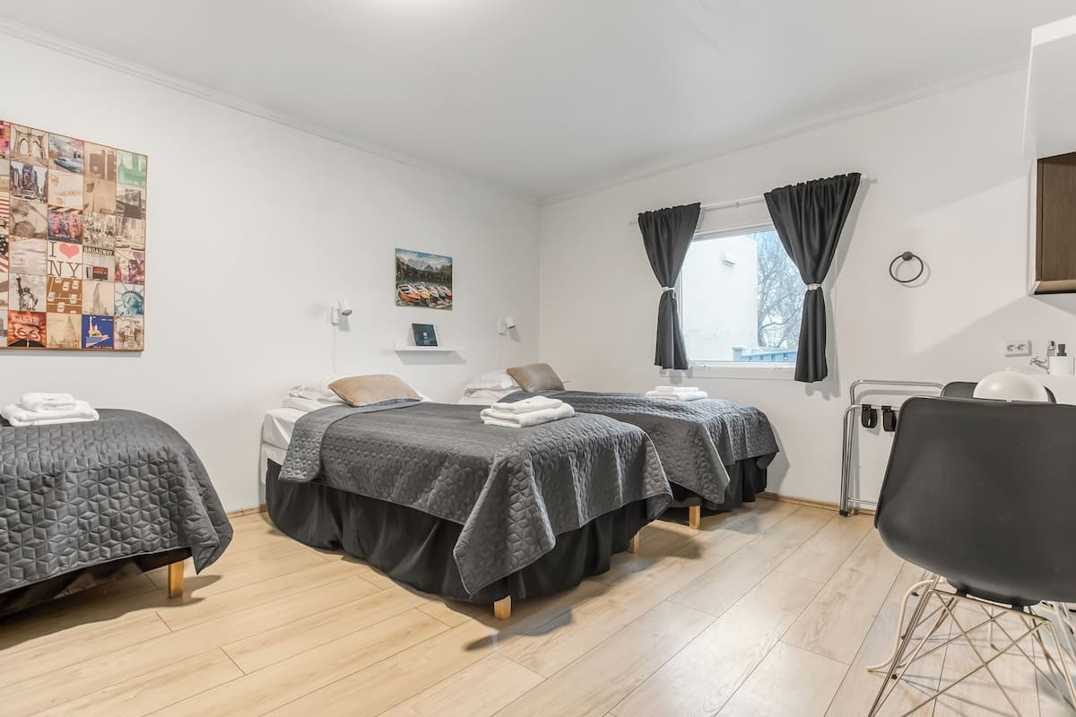 A hotel room with two beds, a desk, a chair, a window with curtains, a wall art piece, and a small shelf with a picture.
