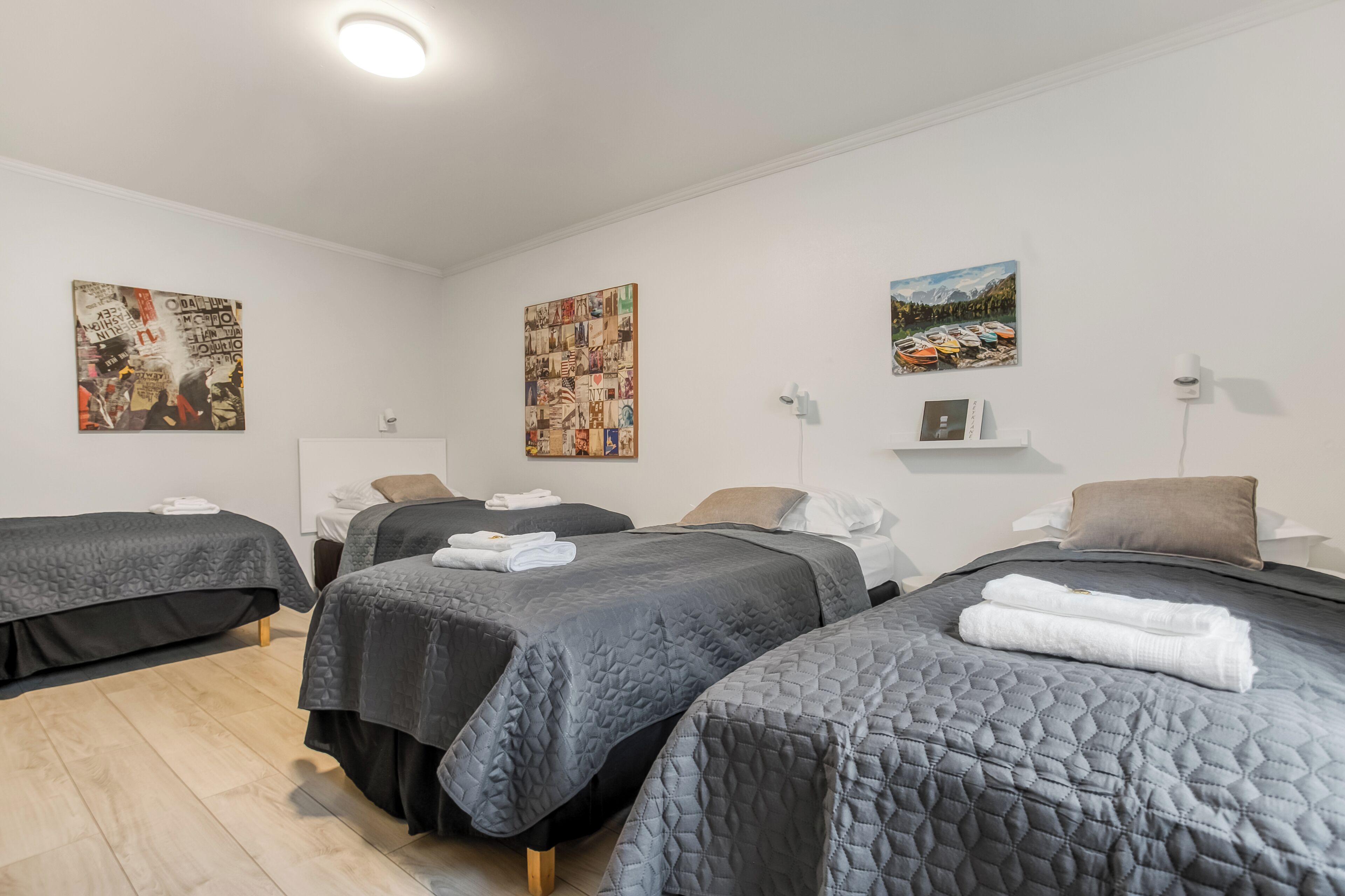 Habitación de hotel con tres camas, cada una con una colcha acolchada oscura y toallas bien dobladas. Se observan obras de arte en la pared y un pequeño estante con un libro.
