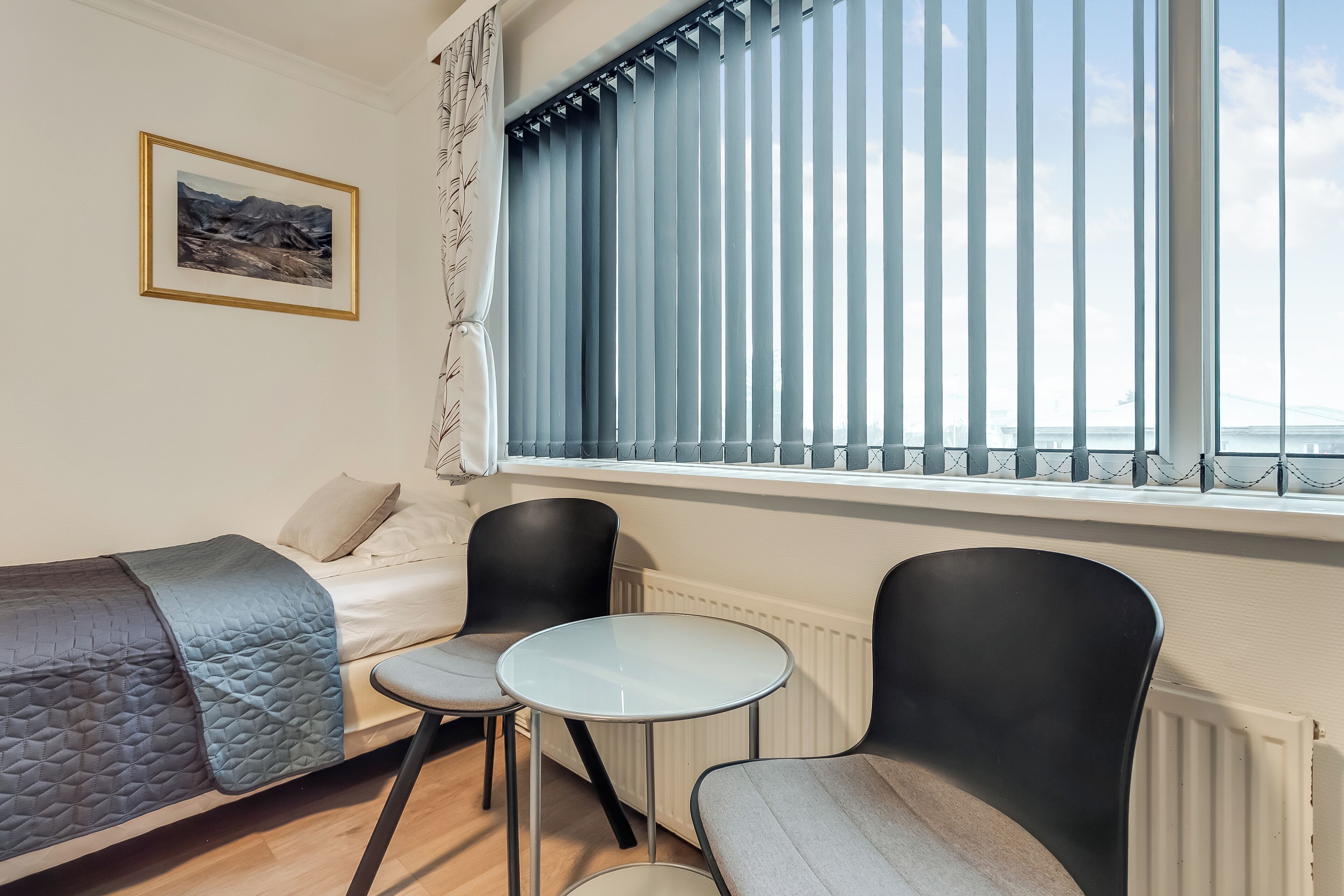Un dormitorio con cama, dos sillas, una mesita, una ventana con persianas, un radiador y un cuadro en la pared.