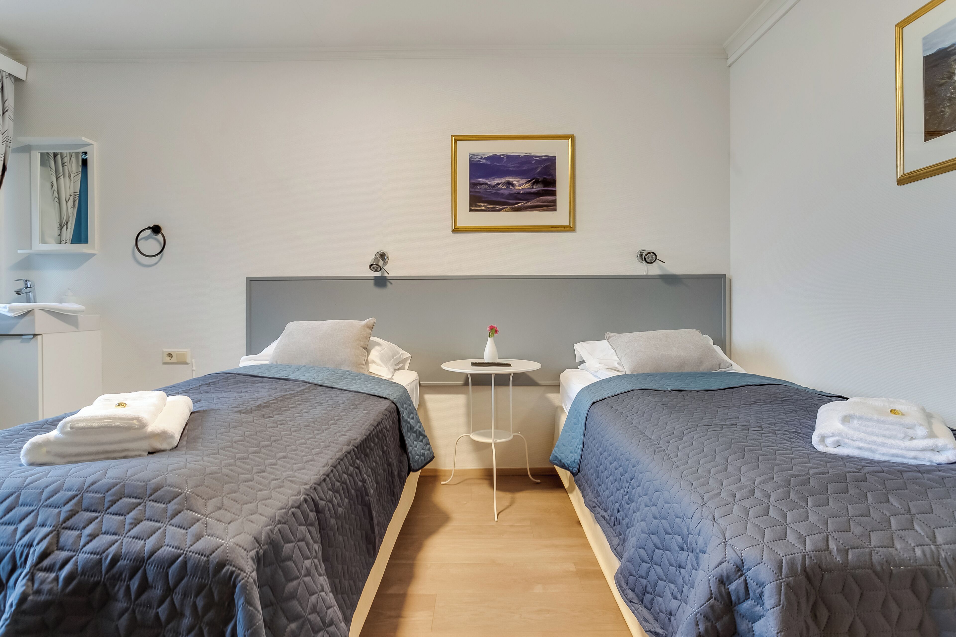 A hotel room with two beds, a small table with a vase, and a painting on the wall.