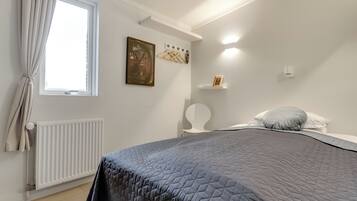 A bedroom with a bed, a radiator, a window with curtains, a wall-mounted shelf, and a wall-mounted light fixture.
