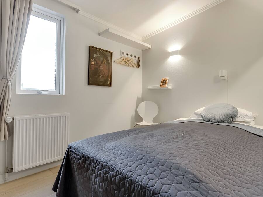 A bedroom with a bed, a radiator, a window with curtains, a wall-mounted shelf, and a wall-mounted light fixture.