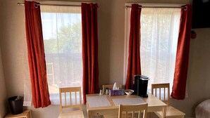 Um quarto com uma mesa e cadeiras de madeira, um banquinho pequeno, uma lixeira e uma janela com cortinas vermelhas.