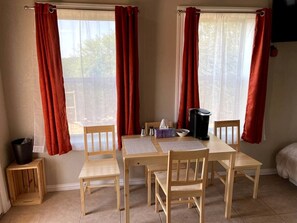 A room with a wooden table and chairs, a small stool, a trash bin, and a window with red curtains.