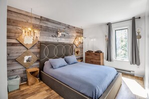 Ein modernes Schlafzimmer mit einem Holz-Kopfteil, einem großen Bett mit blauer Bettdecke, einem Nachttisch aus Holz, einer Kommode und einem Fenster mit Vorhängen.