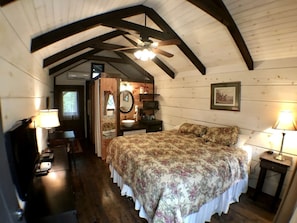 A bedroom with a wooden ceiling, a bed with floral bedding, a bedside table, a mirror, and a window. - Tiny Home Cottage in the Smokies near Sevierville, Tennessee (Sevierville)