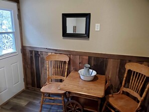 Habitación de esquina con una mesa de madera y dos sillas, un espejo pequeño en la pared y una ventana con persianas.