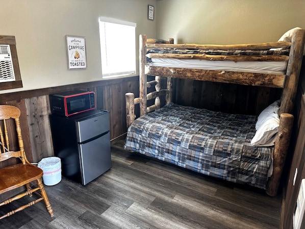 Una cama nido con cubrecama de cuadros, un refrigerador pequeño, un microondas, una silla de madera y un cartel de bienvenida.