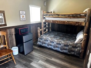 Una cama nido con cubrecama de cuadros, un refrigerador pequeño, un microondas, una silla de madera y un cartel de bienvenida.
