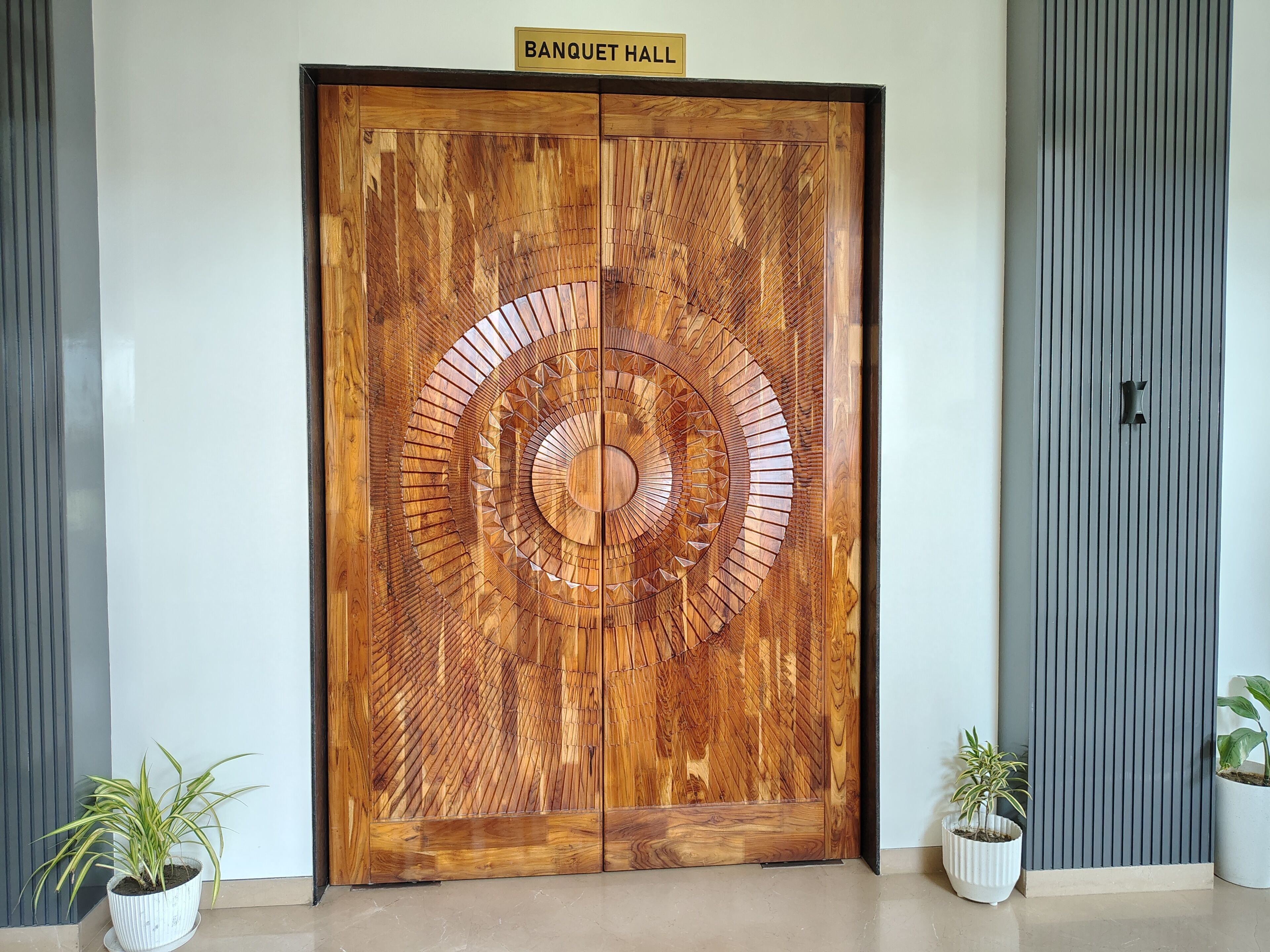 Une grande porte en bois, ornée d’un motif circulaire, portant l’inscription « SALLE DE BANQUET », et flanquée de panneaux verticaux et de plantes en pot.