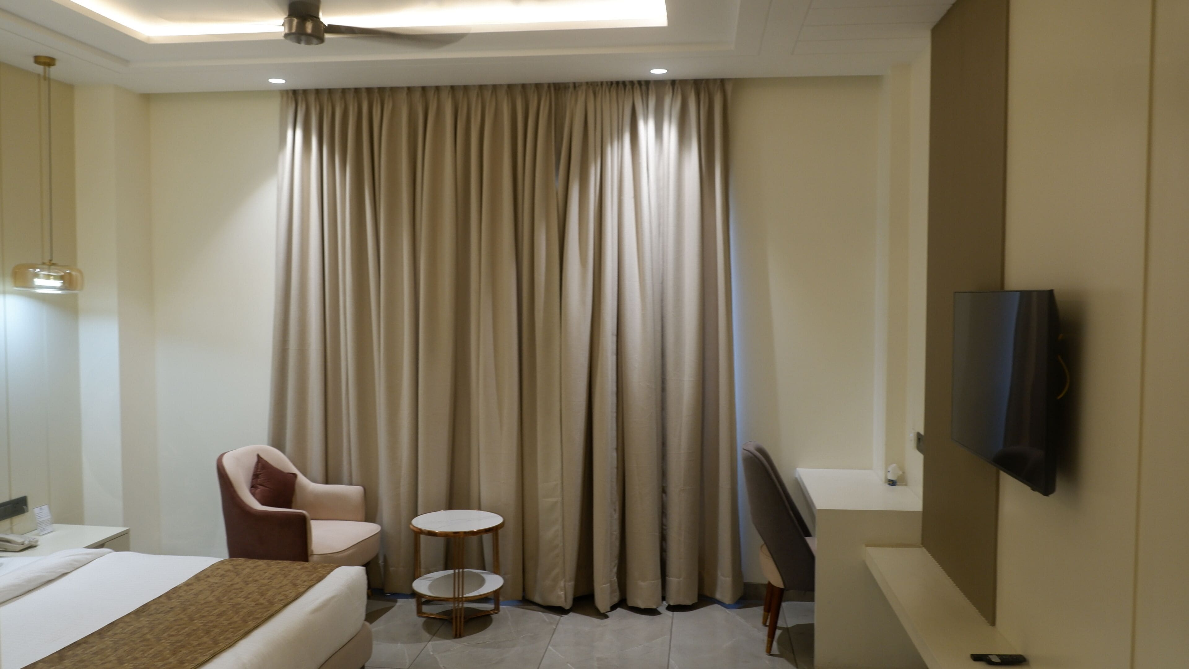 Une chambre d’hôtel avec un lit, une chaise, une petite table, un téléviseur et une grande fenêtre avec des rideaux.