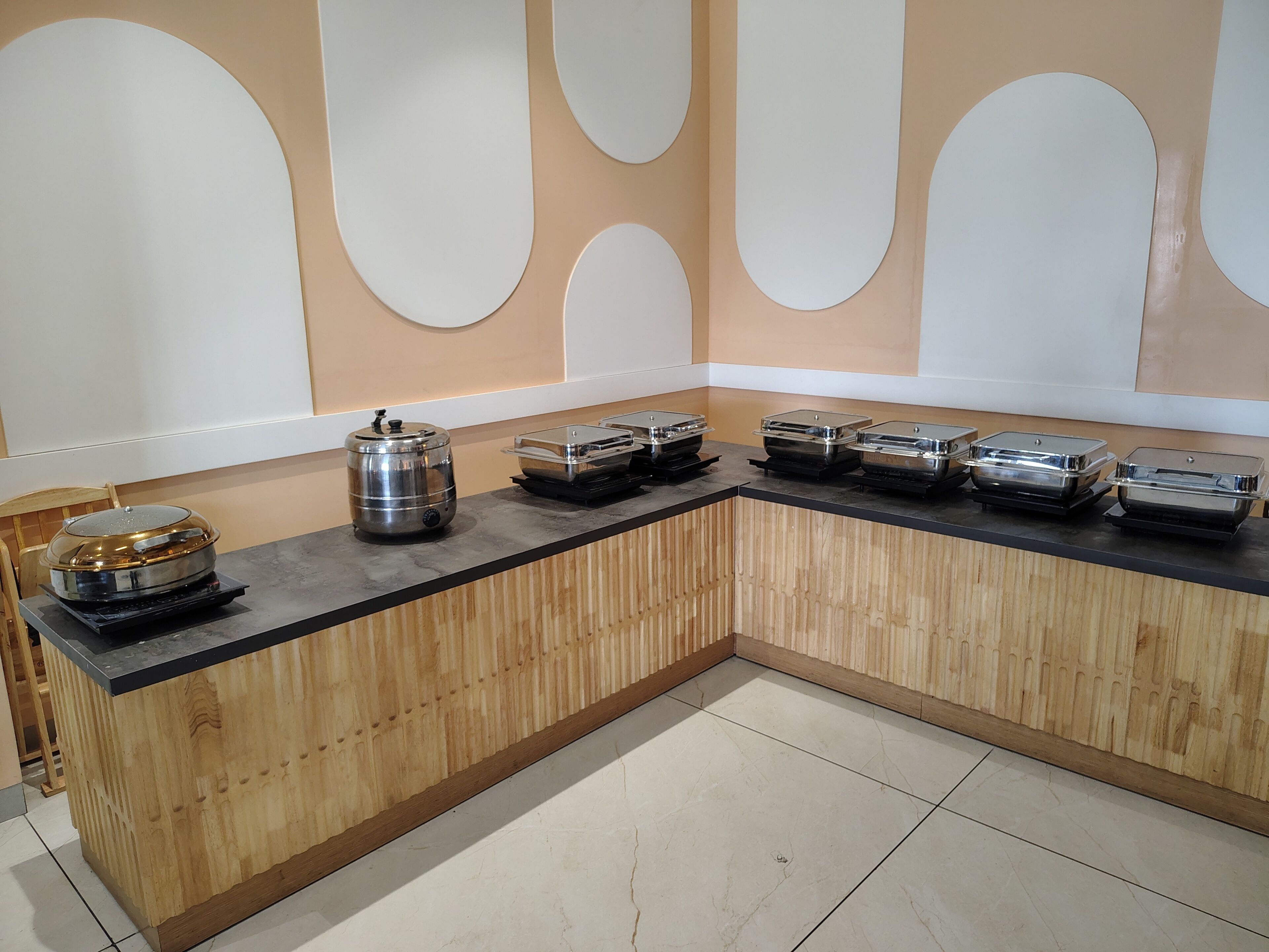A buffet setup with a wooden counter, stainless steel serving dishes, and a tiled floor.