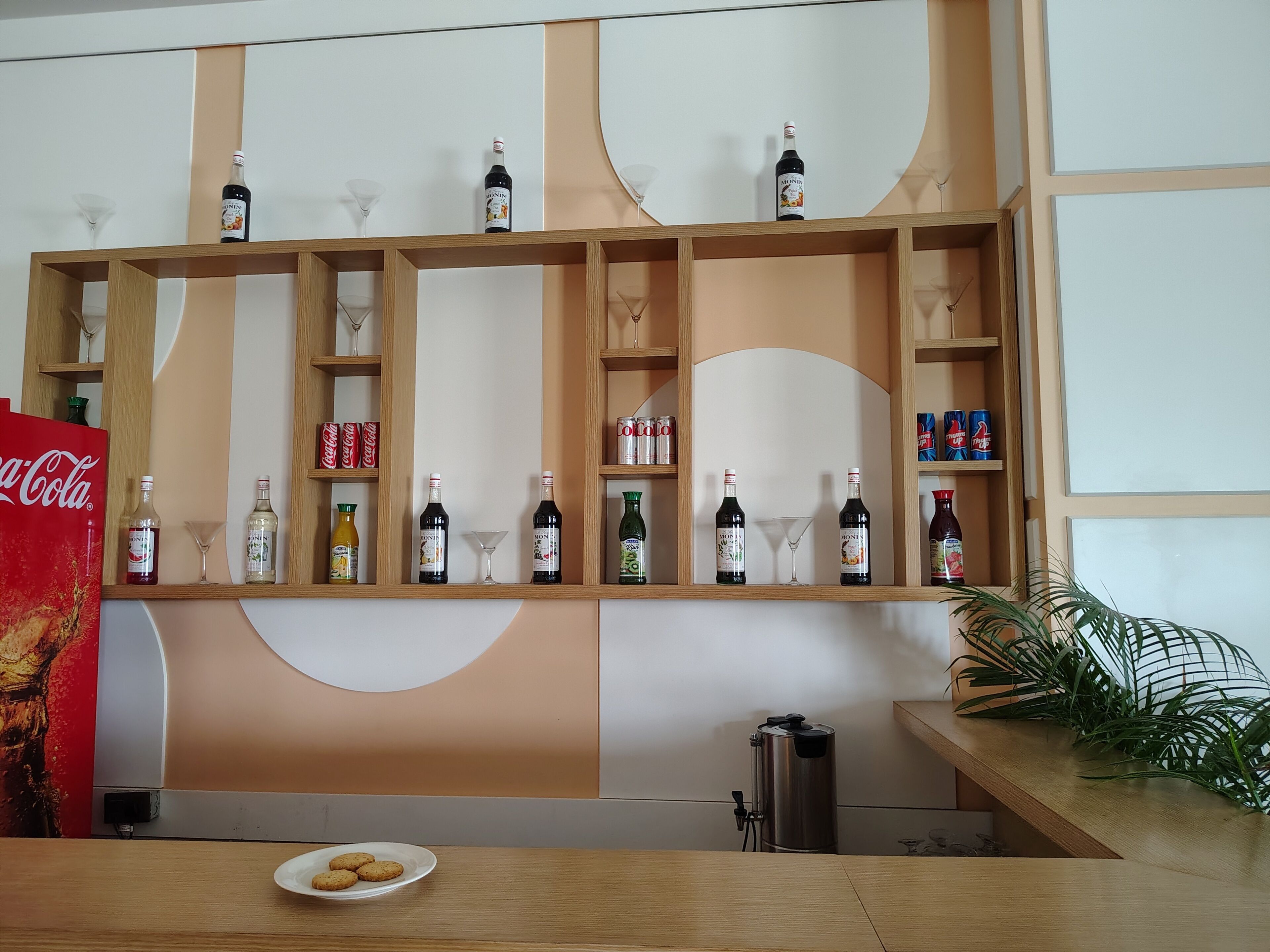 Una zona de bar con estantes llenos de botellas y vasos, un refrigerador de Coca-Cola y un plato de galletas sobre una mesa.