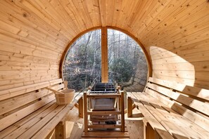 A wooden sauna with benches and a grill, surrounded by a circular wooden structure with a view of trees.