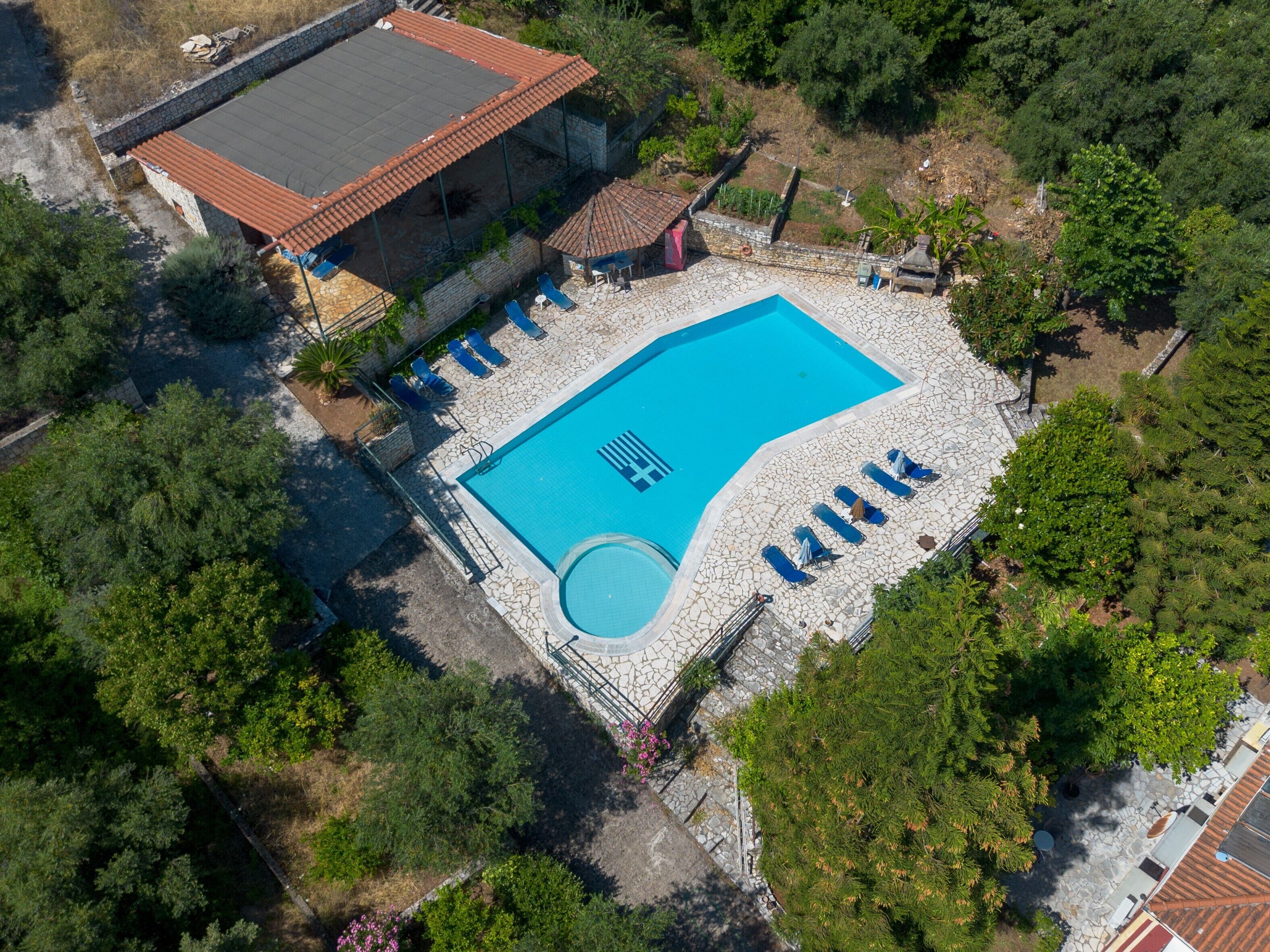 An aerial view of a swimming pool surrounded by stone tiles, with lounge chairs and a covered area.