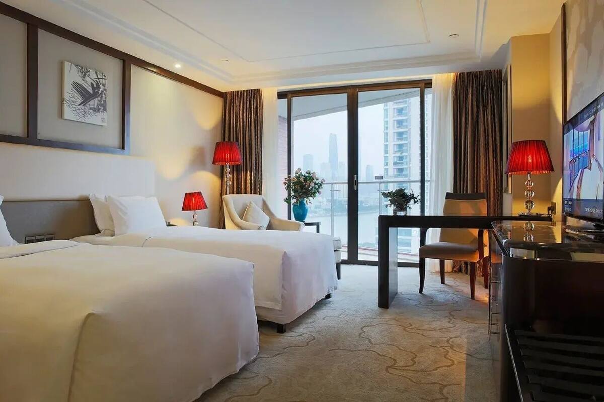 Habitación de hotel con dos camas, un escritorio con computadora, una silla y un ventanal con vista a edificios.