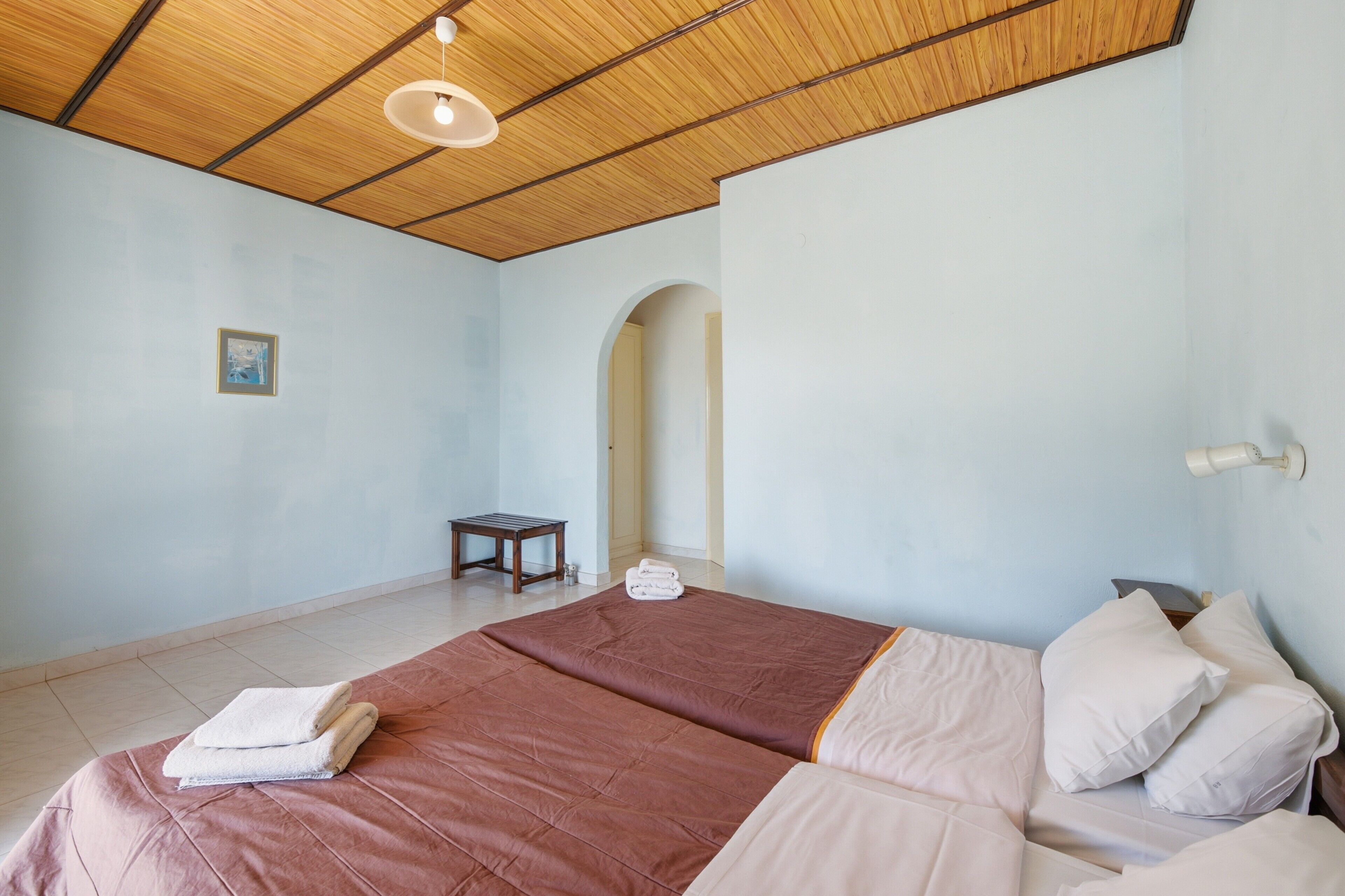 Een slaapkamer met een houten plafond, een bed met twee laken, een klein tafeltje en een wandlamp.