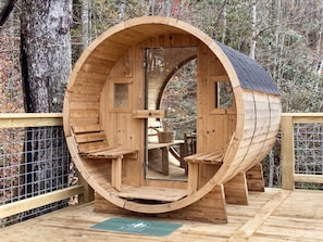 Una estructura de madera con forma de barril, con una puerta, un banco en su interior y una terraza de madera en el exterior.
