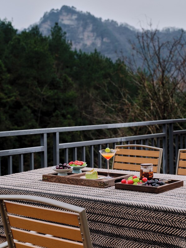 Una mesa con pastel, frutas, una bebida y una jarra en un balcón con sillas y vistas a las montañas.