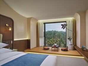 Una habitación de hotel con un ventanal grande, una cama, una mesita de noche, una lámpara y vistas a un área boscosa.