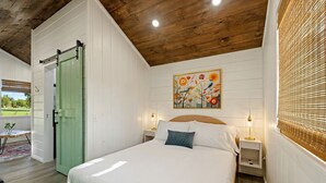 Un dormitorio con cama, mesitas de noche, una puerta verde, un cuadro y una ventana con persianas.