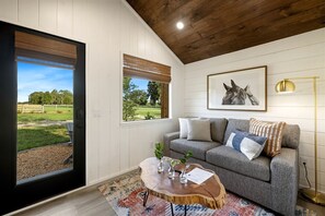 Ein Zimmer mit grauem Sofa, einem Holztisch, einem gerahmten Bild von einem Pferd und Blick auf eine grüne Fl äche durch eine Tür.