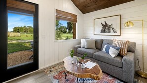 Une pièce avec un canapé gris, une table basse en bois, un tableau représentant un cheval encadré, et une vue sur un espace verdoyant par une porte.