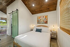 Ein Schlafzimmer mit einem Bett, Nachttischen, einer grünen Tür, einem Gemälde und einem Fenster mit Jalousien.