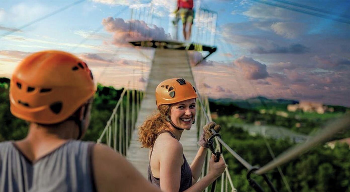 A person wearing a helmet and holding a rope, standing on a wooden bridge with a scenic landscape in the background.
