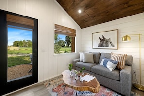 A room with a grey sofa, a wooden coffee table, a framed horse picture, and a view of a grassy area through a door.