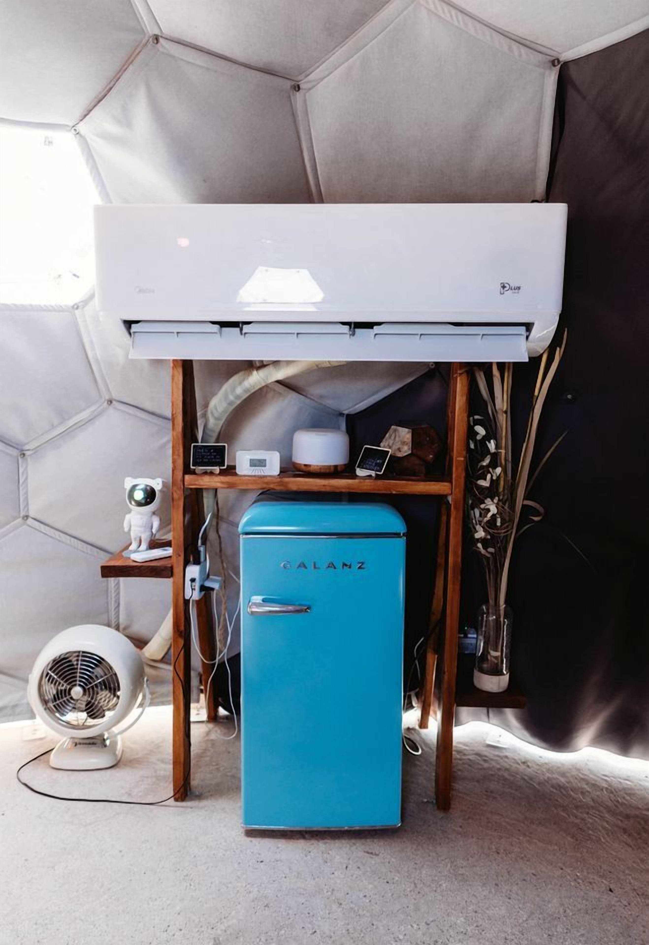 A compact room with a blue refrigerator, a white air conditioner, and a small shelf with a clock and a mobile phone.