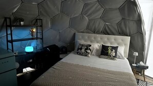 Una stanza con soffitto esagonale, una testiera trapuntata, un letto con cuscini a fantasia, una scrivania con lampada e una piccola pianta.