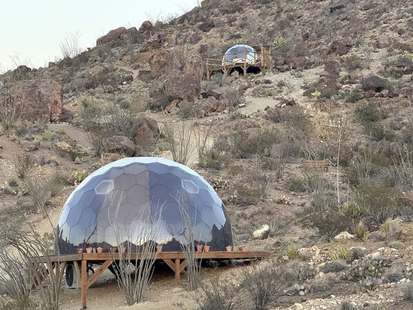 A dome-shaped structure with a reflective surface, situated on a wooden platform in a desert environment with sparse vegetation and rocky terrain.