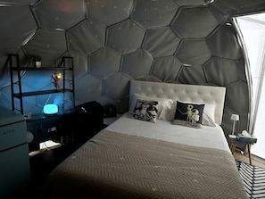 A room with a hexagonal ceiling, a tufted headboard, a bed with patterned pillows, a desk with a lamp, and a small plant.