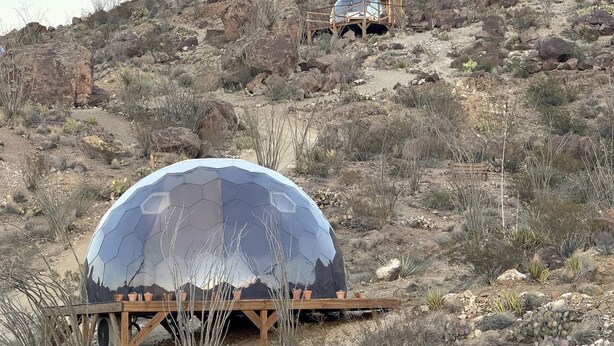 Una struttura a forma di cupola con superficie riflettente, posizionata su una piattaforma di legno in un ambiente desertico con vegetazione rada e terreno roccioso.