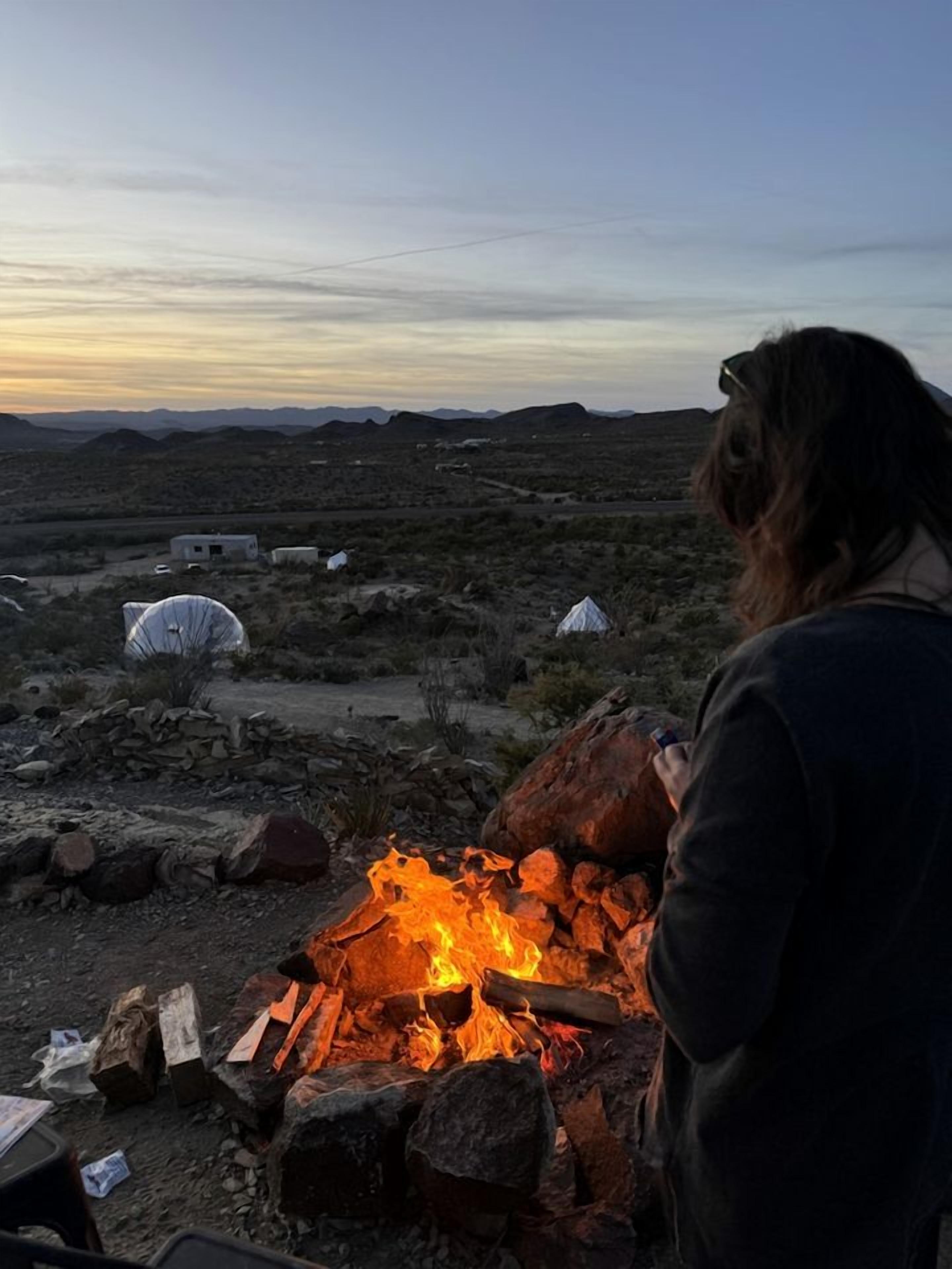 Paisaje desértico con fogata, tiendas de campaña y una persona de pie junto al fuego.