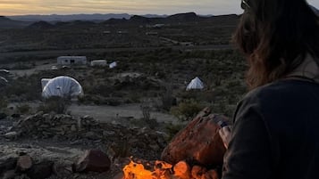 Paisaje desértico con fogata, tiendas de campaña y una persona de pie junto al fuego.