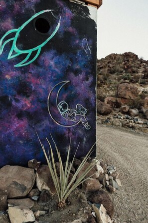 A wall with space-themed graffiti, including a crescent moon and a rocket, and a rocky terrain with sparse vegetation.