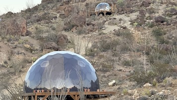 Une structure en forme de dôme, dotée d’une surface réfléchissante, qui se trouve sur une plateforme en bois, dans un environnement désertique caractérisé par une végétation clairsemée et un terrain rocheux.