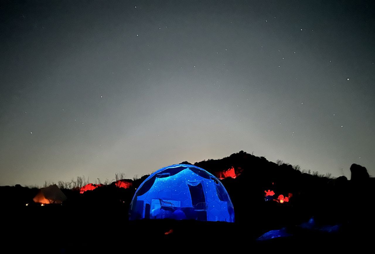 A dome-shaped structure illuminated in blue, surrounded by a dark landscape with sparse vegetation.