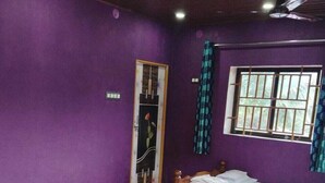 A room with purple walls, a wooden bed, a wooden table, and a window with bars. - Naga Chowdeshwari Homestay (Kumta)