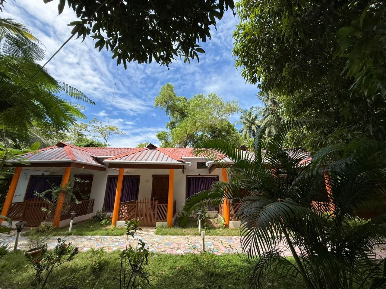 A two-story house with a red roof and orange pillars, surrounded by lush greenery and a patterned walkway.
