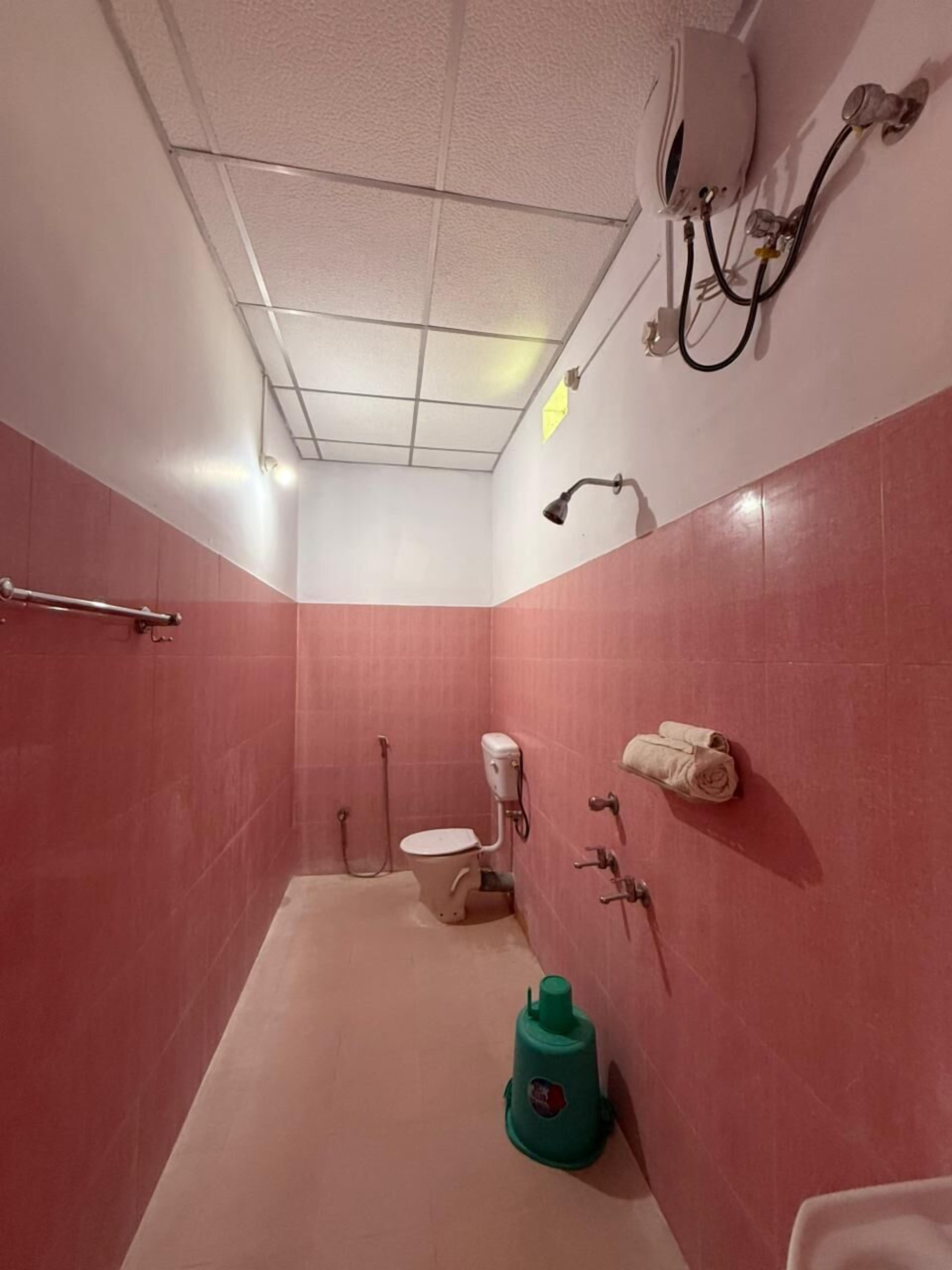 Un baño pequeño y cerrado con azulejos rosas, un inodoro, una ducha de mano, un perchero para toallas y un dispensador de agua verde.