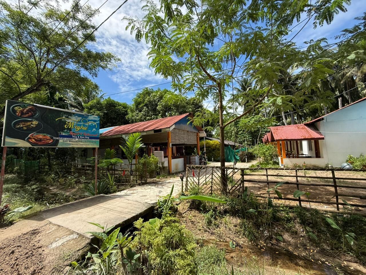 A rural setting with a small building that appears to be a restaurant or cafe, a dirt path, and a signboard advertising food.