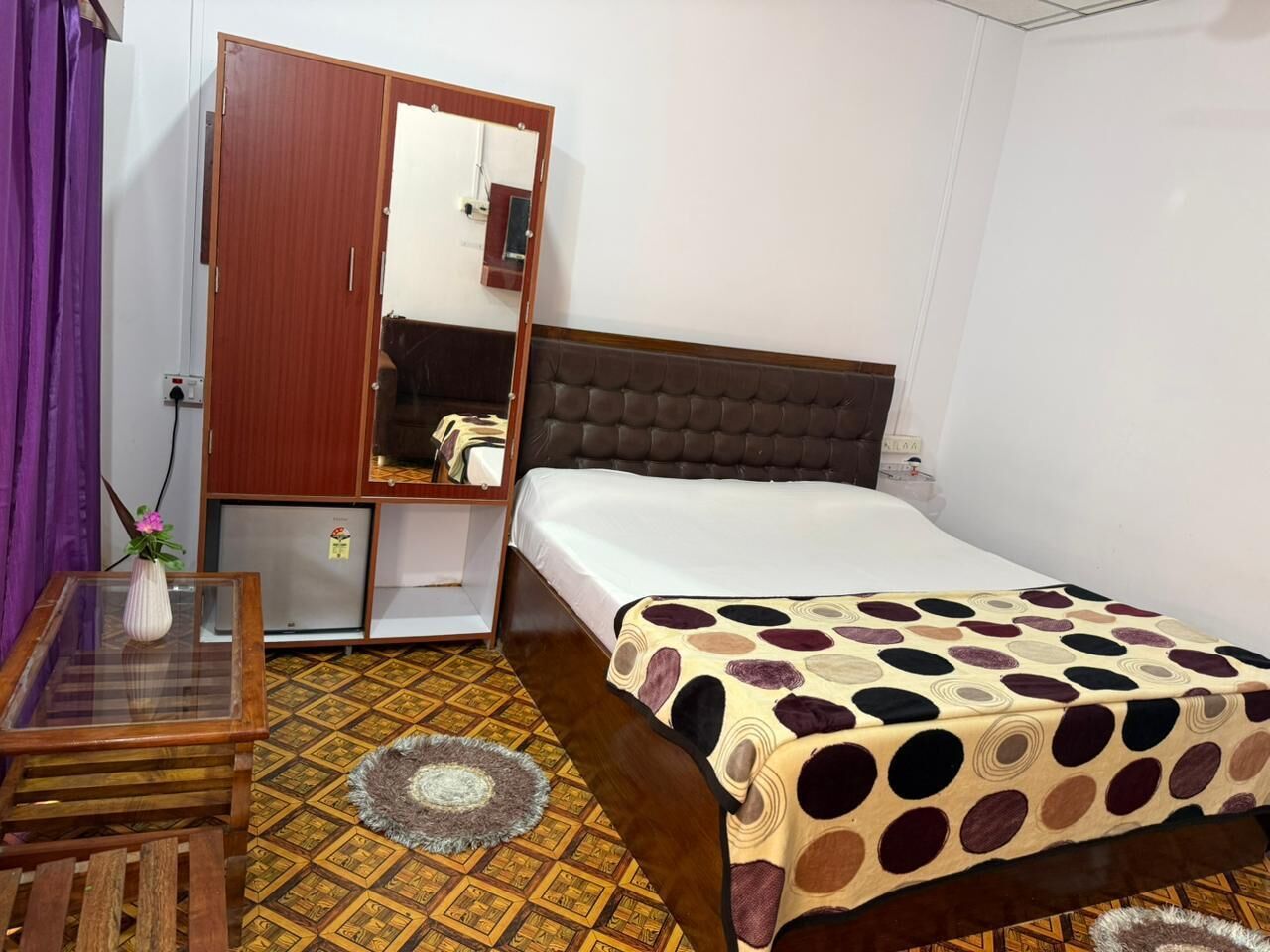 A bedroom with a bed, a wardrobe, a small table with a vase, and a patterned rug.