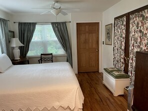 Une chambre à coucher comprenant un lit, une chaise, un bureau, une fenêtre avec des rideaux, une porte et un ventilateur de plafond.